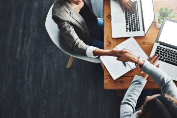 Two professionals shaking hands across a desk with laptops and notes.
