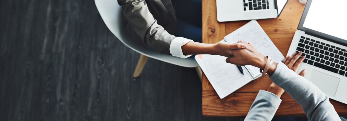 Two professionals shaking hands across a desk with laptops and notes.