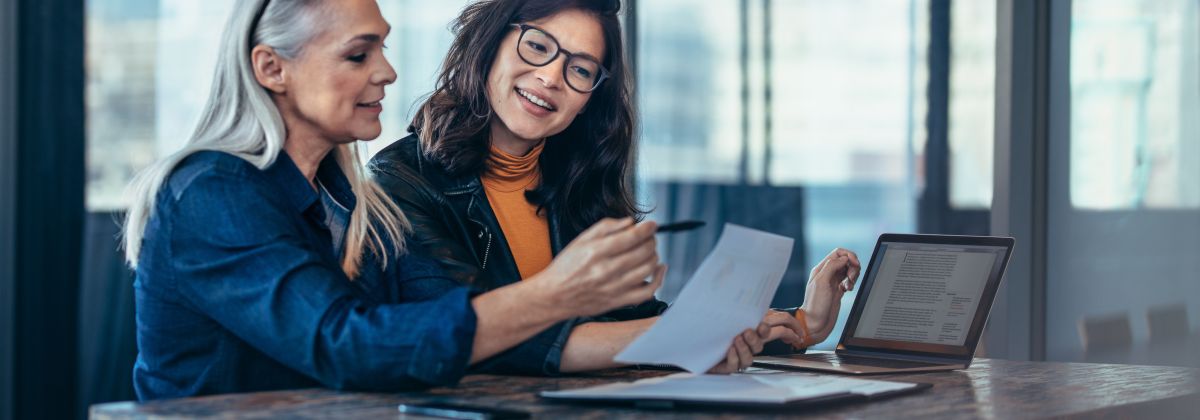 Two females in corporate environment reviewing printed and digital documents. 